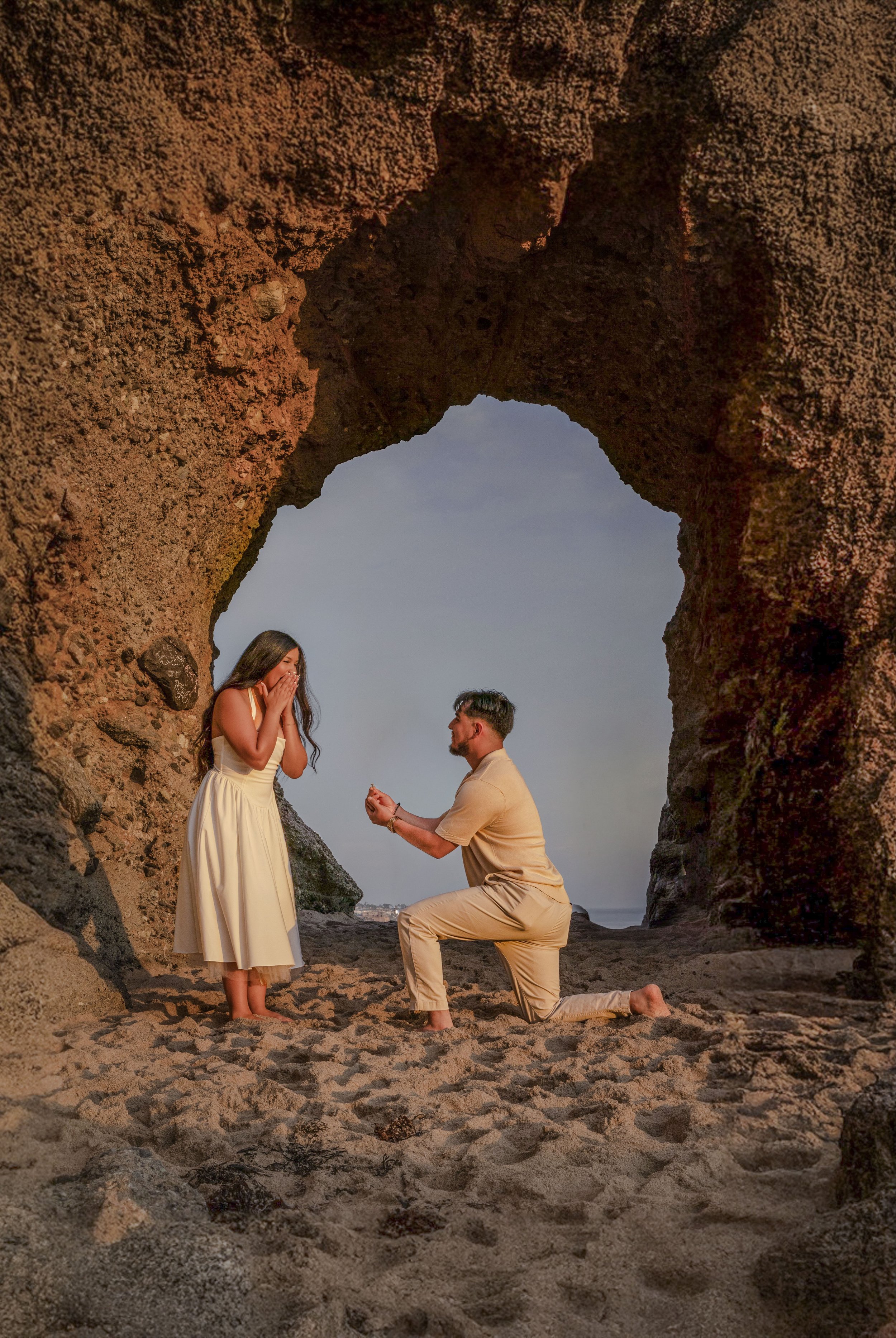 A man proposing marriage to a woman on a beach during sunset, framed by a natural rock formation with a heart-shaped opening.