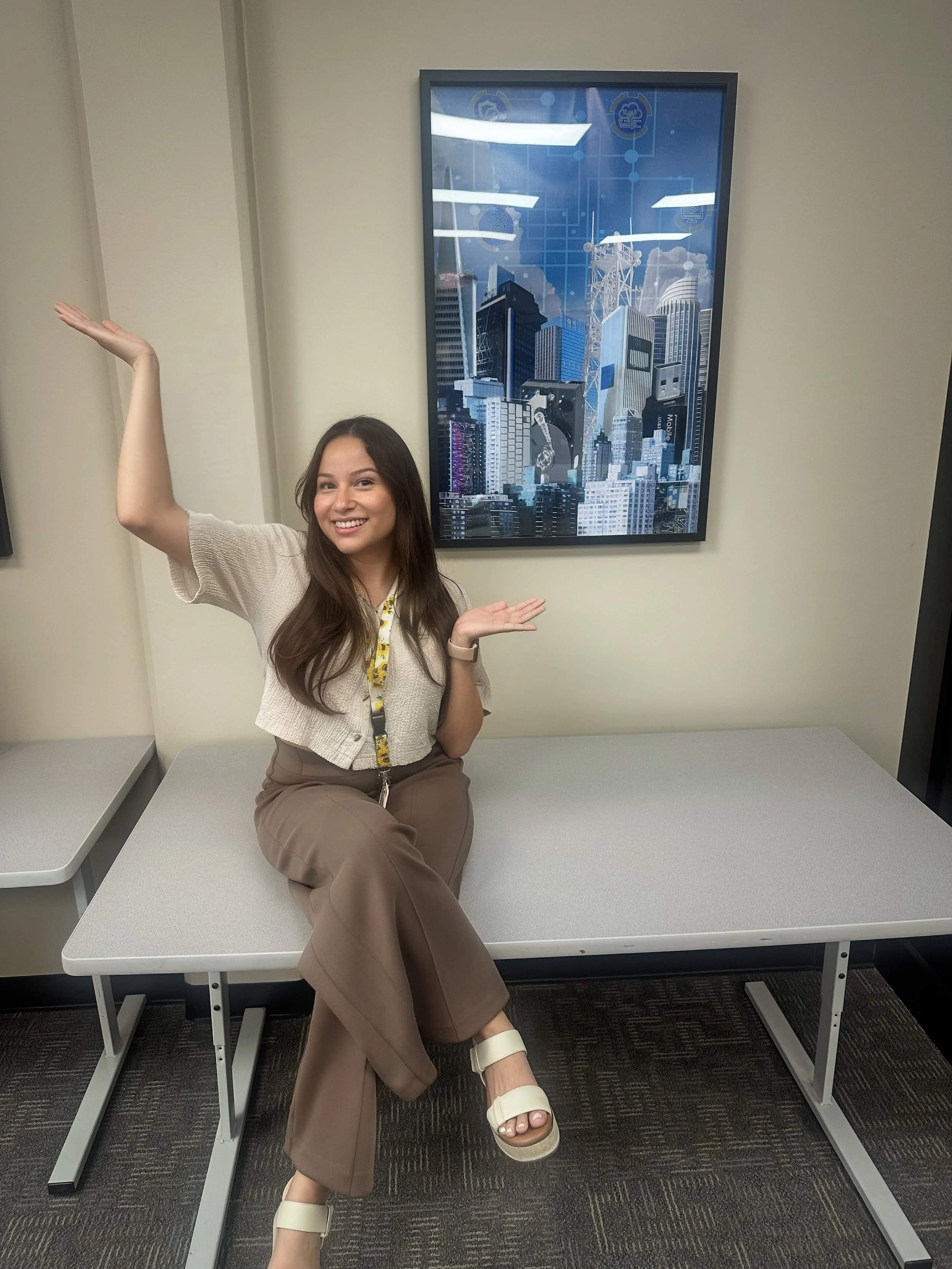 A woman sitting on a gray table in an office, smiling, with her arms raised and hands open, with a city skyline with tall buildings and a blue sky in a framed picture on the wall behind her.