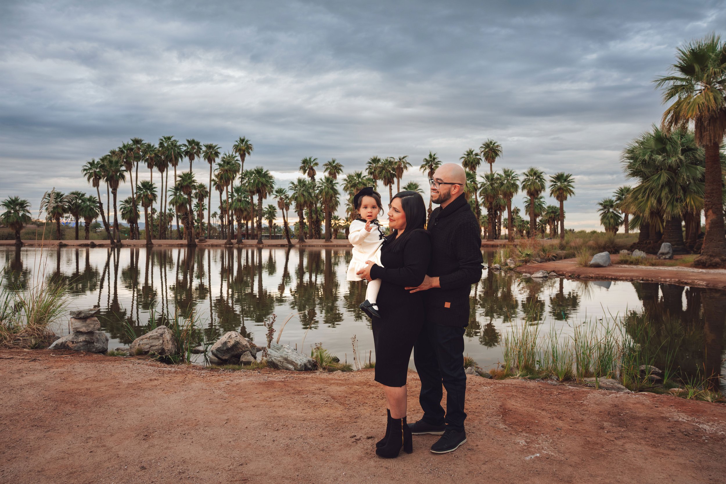 A family of three standing by a lake with palm trees in the background during cloudy weather.