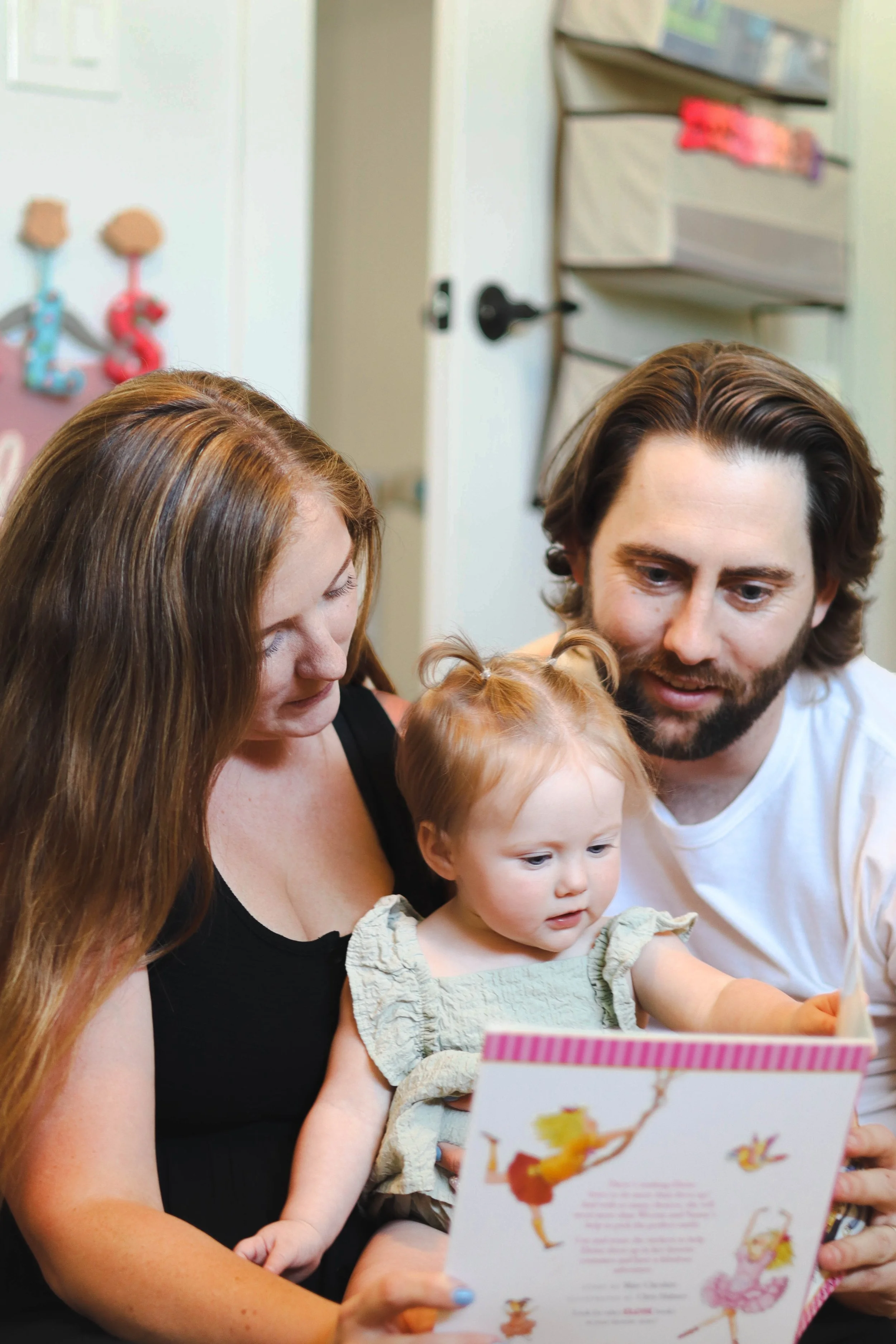 A family of three, including a woman, a man, and a young girl, sit closely together reading a colorful children's book.