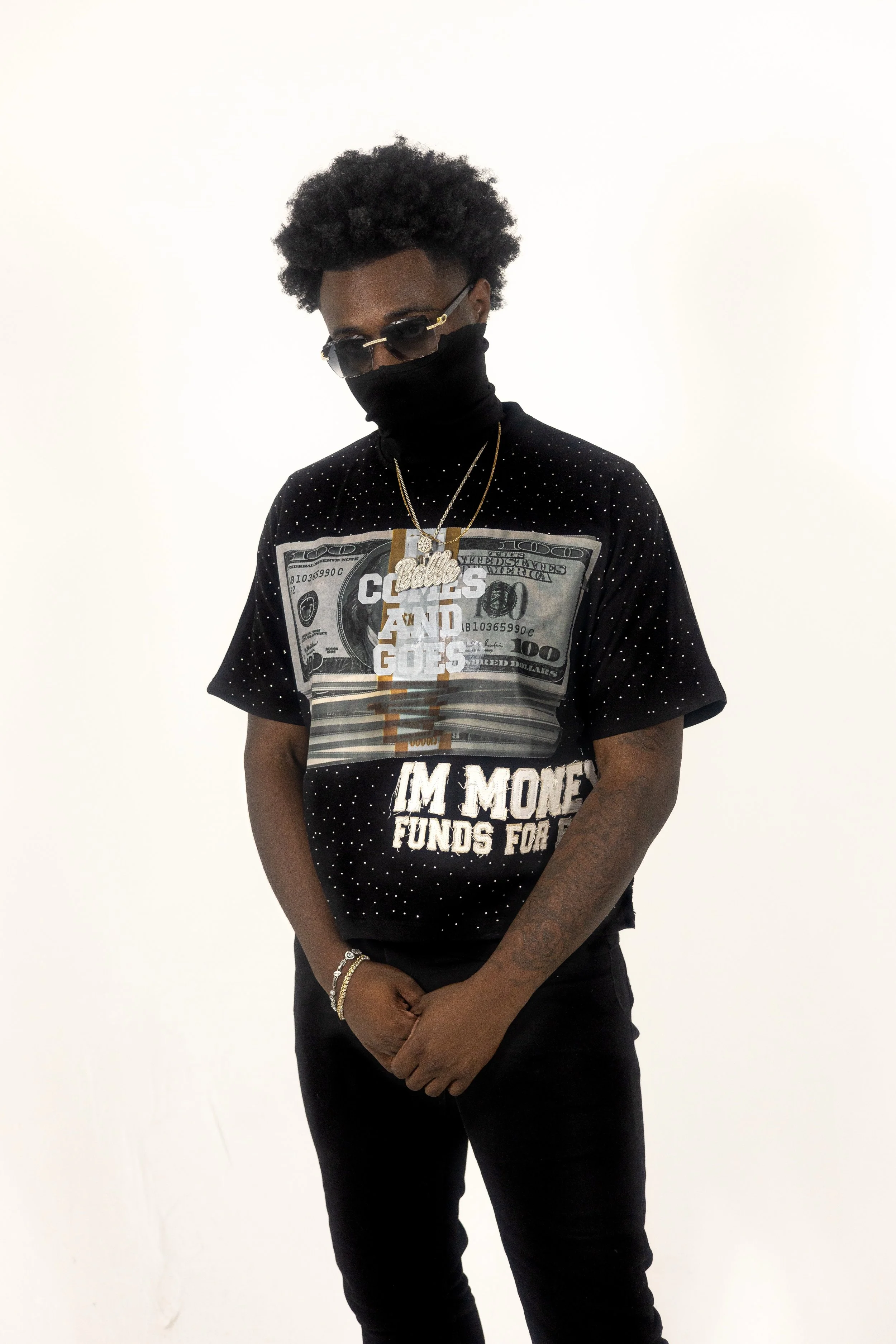 Person wearing a black shirt with a large money graphic, sunglasses, gold chains, and a black face covering.