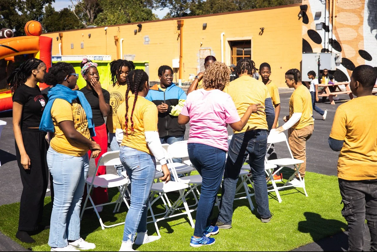Group of people gathering outdoors near white folding chairs and an inflatable bounce house.