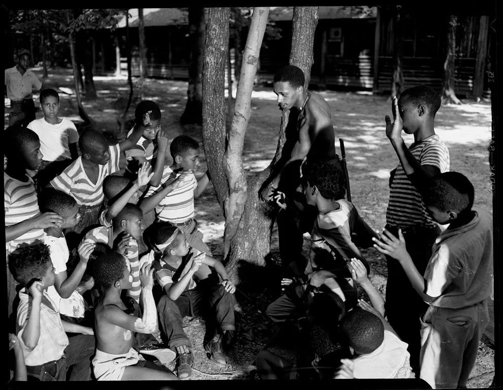 A group of African American children gathered outdoors around a young man, who is leaning against a tree, teaching or telling a story. The children are sitting and raising their hands in interaction. The scene is in a wooded area with cabins in the background.