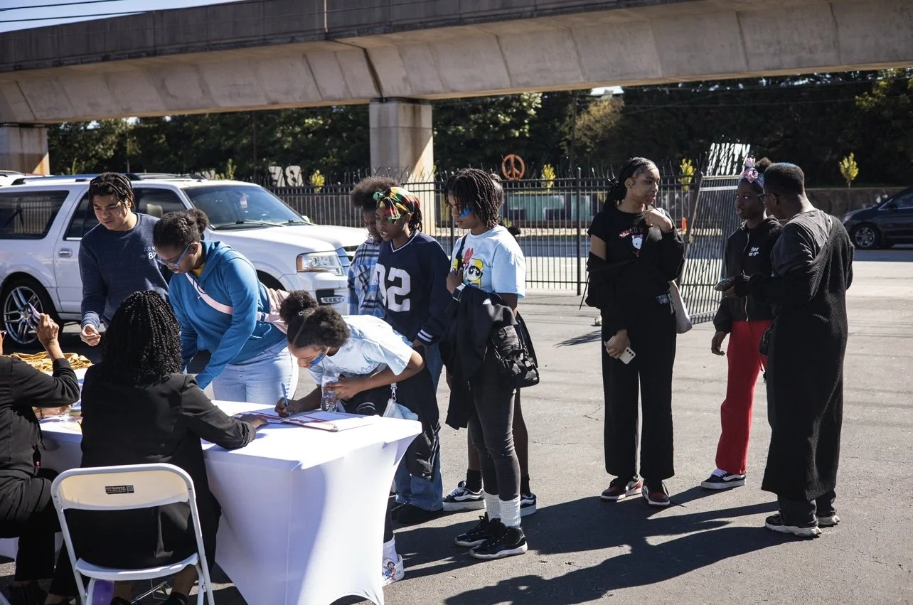 Group of people gathered outdoors, some standing and others sitting at a table with paperwork, near parked vehicles under a bridge.