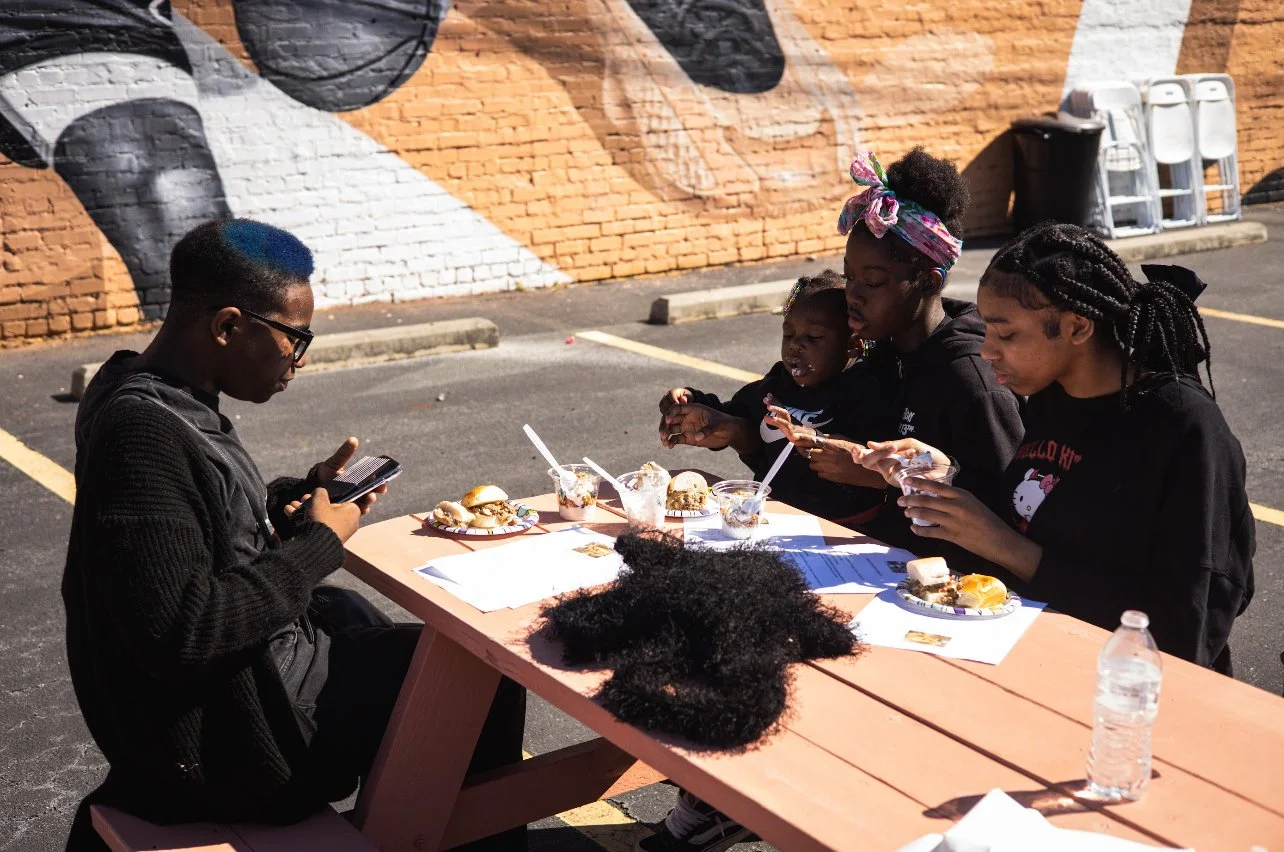 Group of people sitting at a picnic table outdoors, eating food and using a smartphone; mural visible in the background.