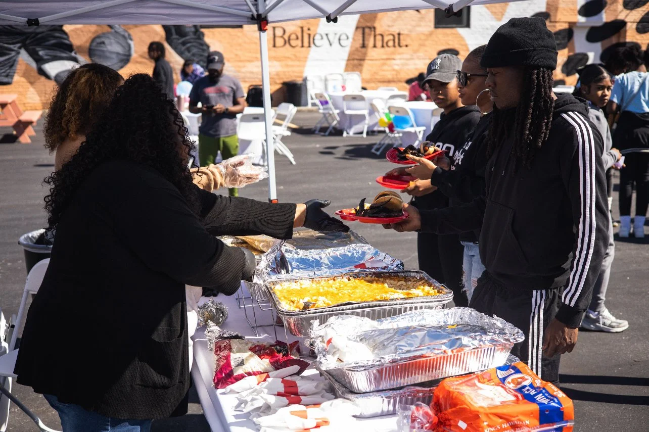 People serving food at an outdoor event, with trays of food and bread on a table, attendees holding red plates waiting in line.