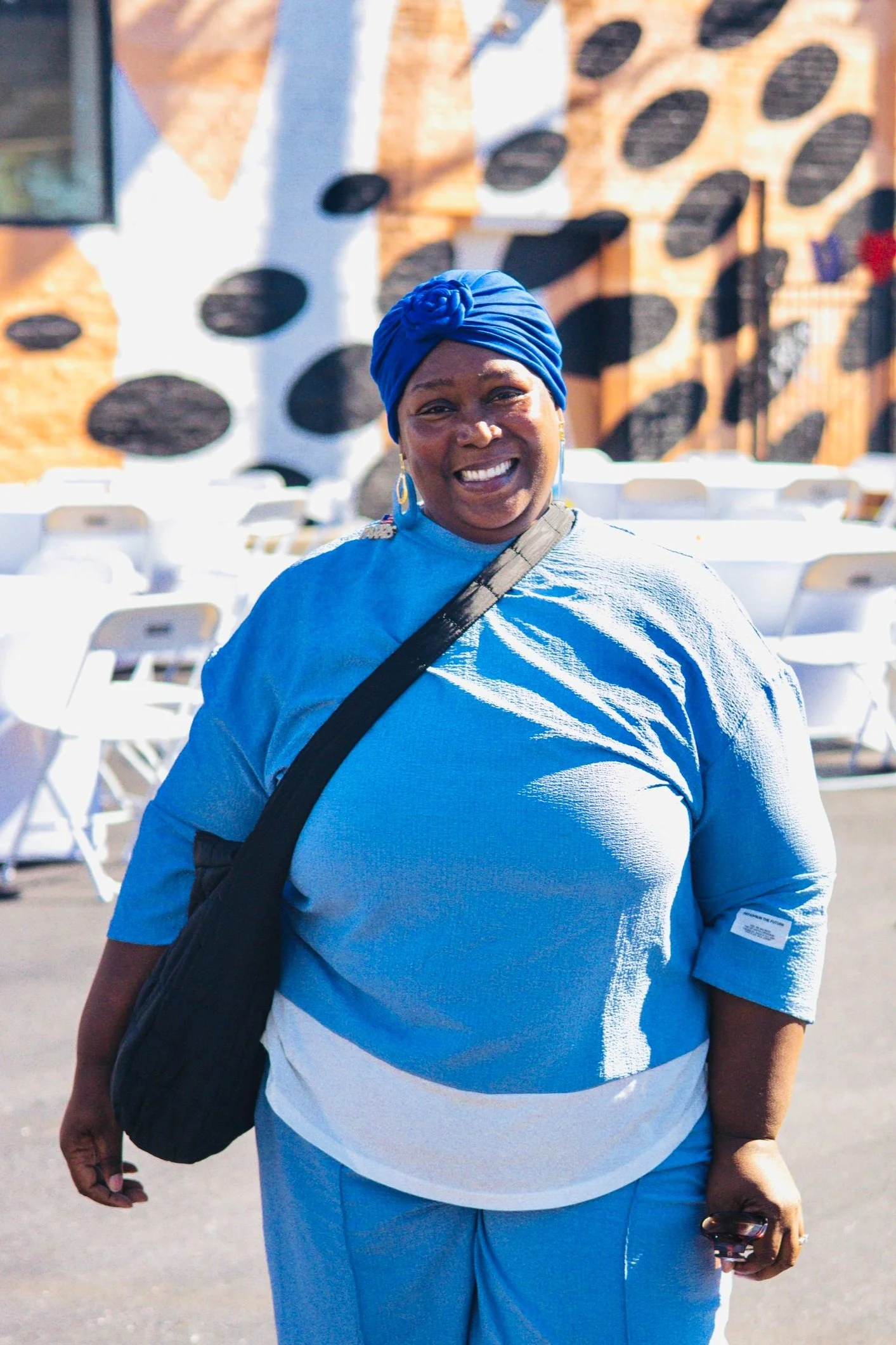 Person in blue outfit with headwrap smiling outdoors, colorful wall in background.