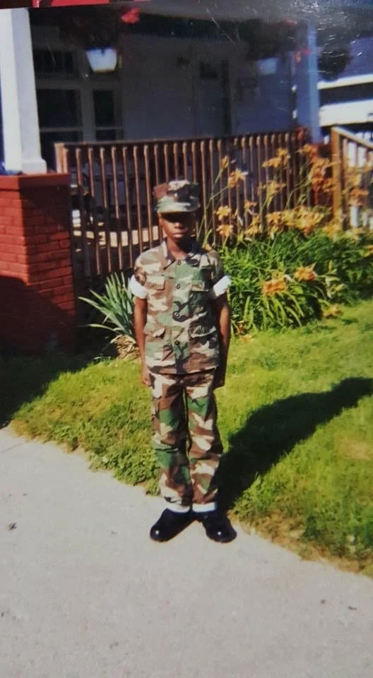 Person in military uniform standing on a sidewalk in front of a house with a porch and garden.