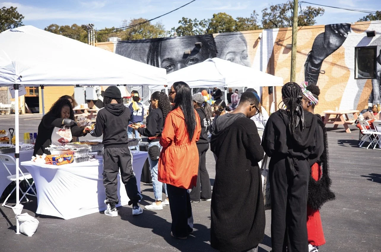 People gathered at an outdoor food event with white tents and a mural on a building wall.