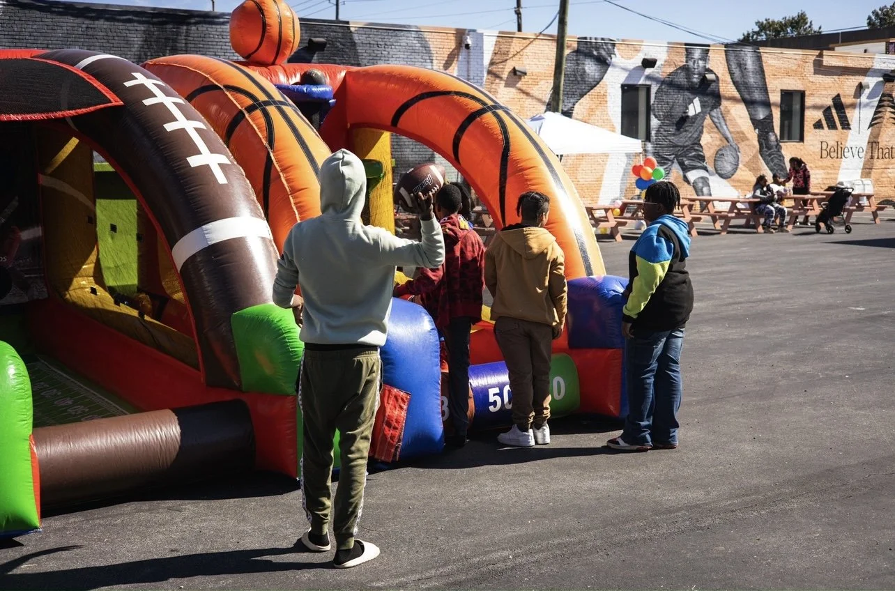 People gathered around an inflatable sports-themed game with football and basketball designs, outdoors on a sunny day.