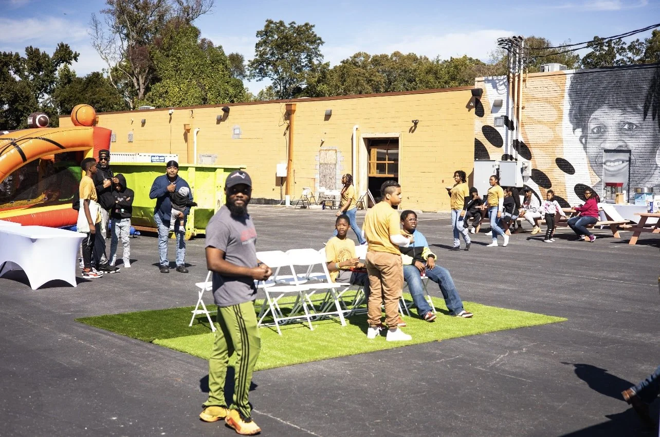 Outdoor community gathering with people sitting and standing around, inflatable play structure, and mural on a building wall.