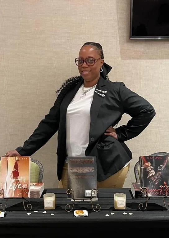 A person stands behind a table displaying books and candles. The person is wearing glasses, a black blazer, and a white shirt.