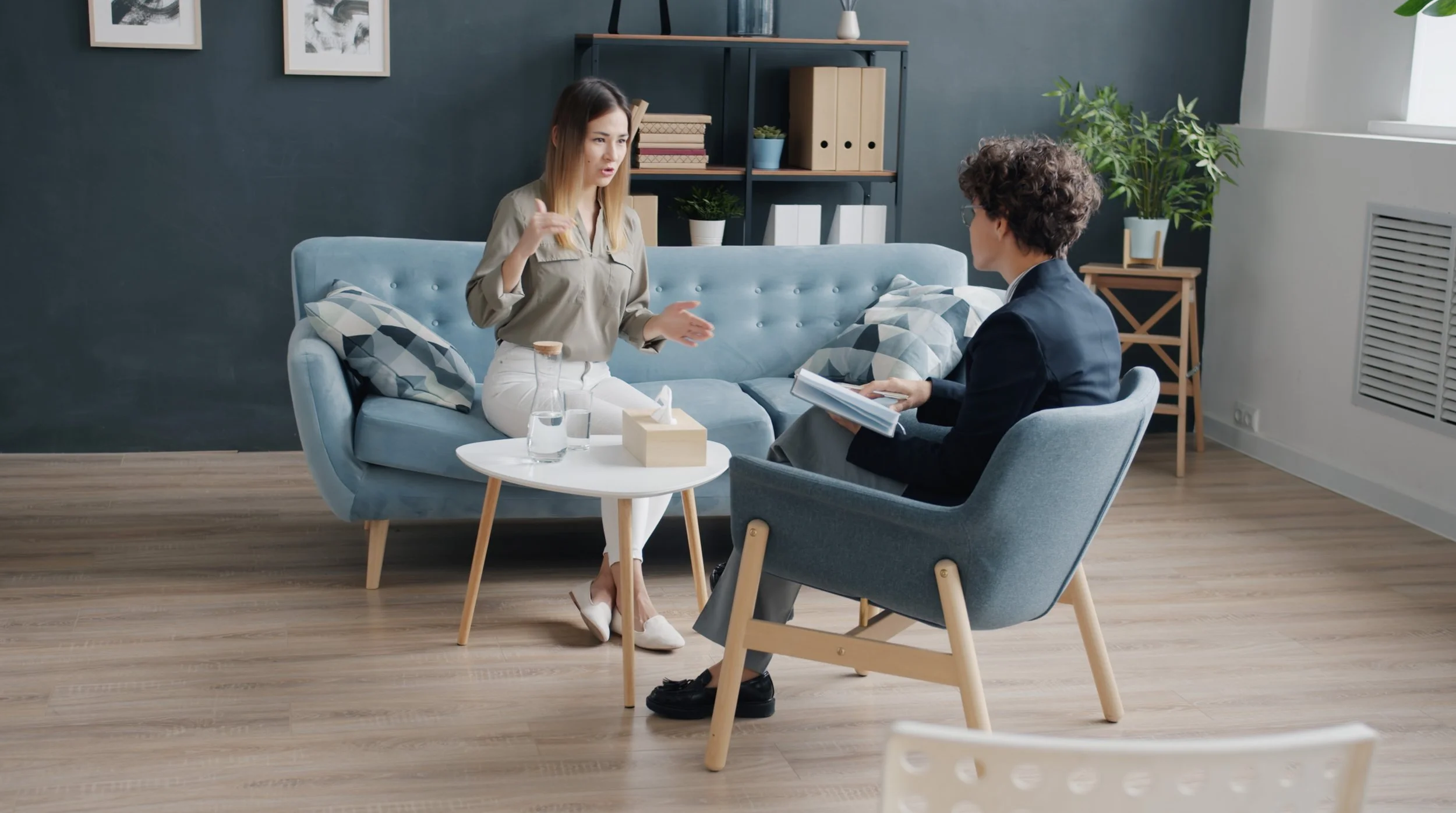 A woman and a man are sitting in a living room having a discussion. The woman is speaking, using hand gestures, while the man is listening and holding a notebook. There is a white coffee table with water bottles and tissue box between them, and a blue sofa and armchair. The room has a dark blue wall, some shelves with books, plants, and decorations, and daylight coming through a window.