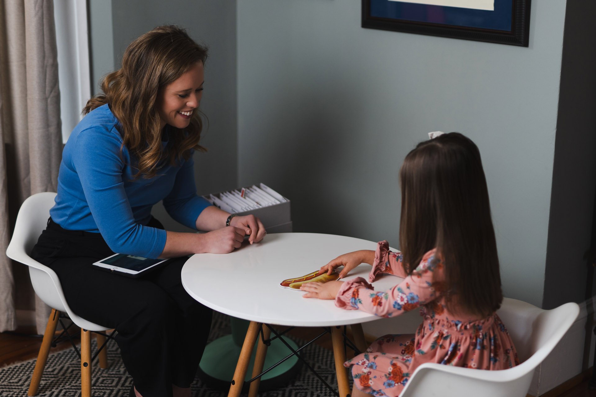 A woman sitting at a white round table smiling and interacting with a young girl as she touches a colorful shape on the table; the woman has light skin, brown hair, and is wearing a blue shirt and black pants, with a tablet on her lap, and the girl h