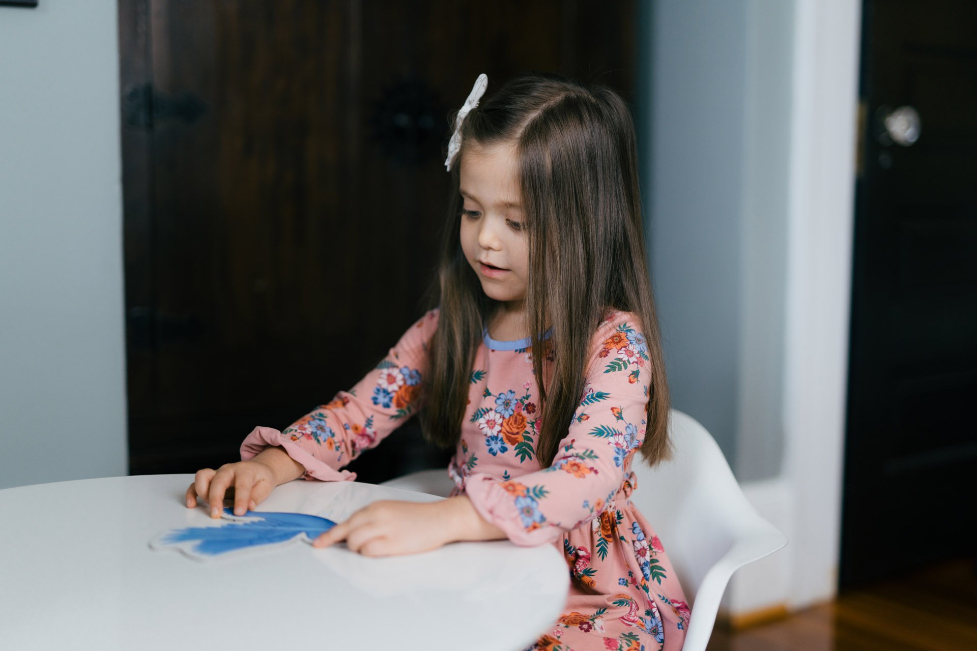 A young girl with long brown hair, wearing a pink dress with floral patterns, is sitting at a white table. She appears to be playing with a puzzle or a piece of paper, with her hands on the table and a focused expression.