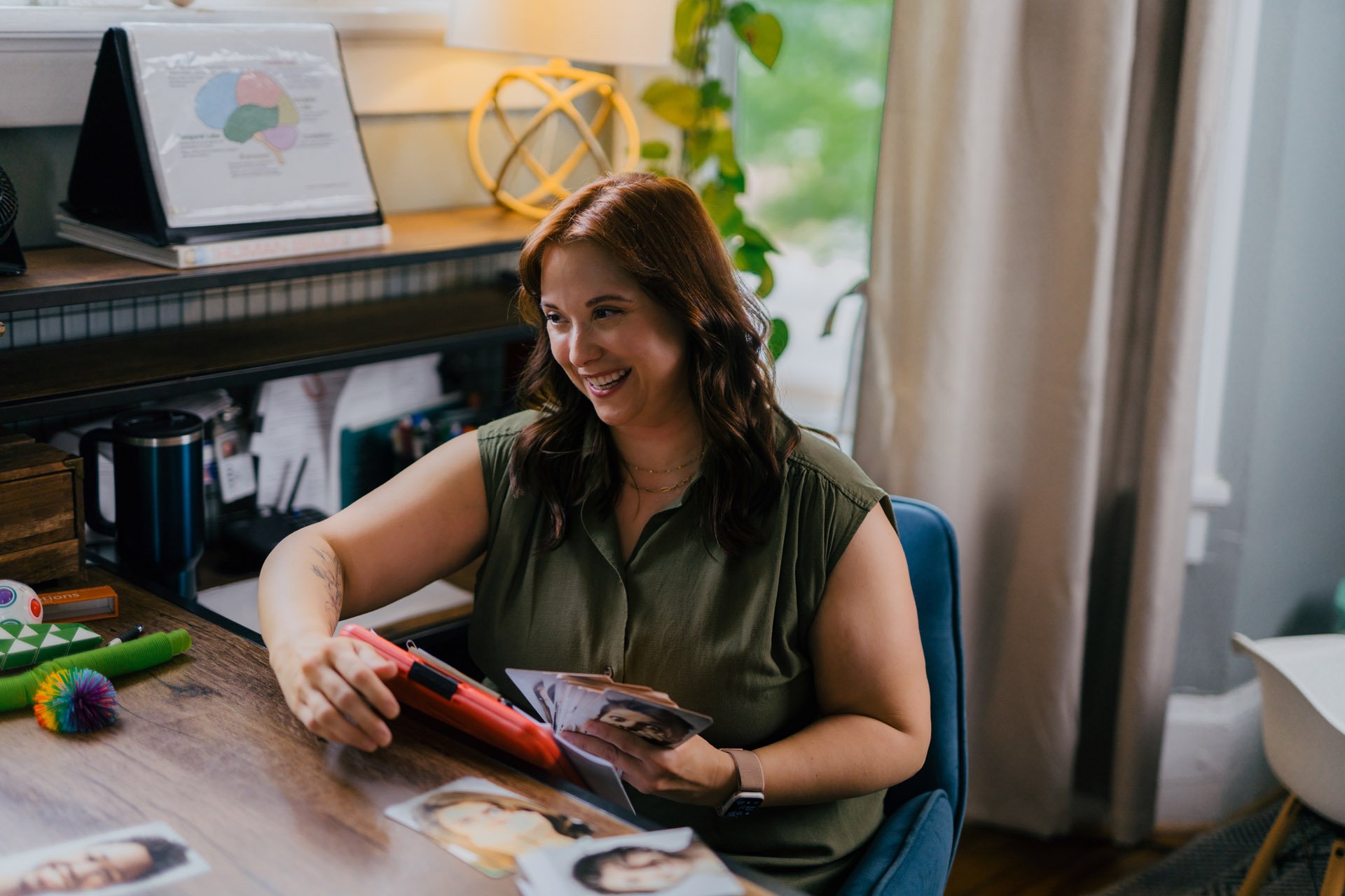 A woman with red hair smiling and looking at photos while sitting at a desk in a home office.