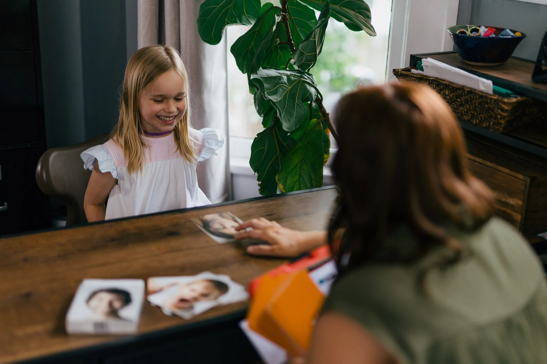 A young girl with blonde hair smiling at a woman across a wooden table, with photos and magazines on the table, inside a room with green plants.