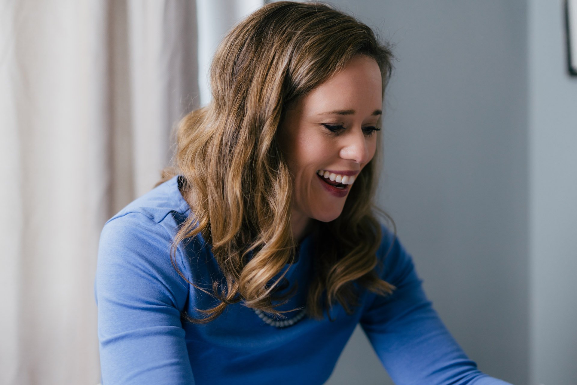 A woman with wavy brown hair smiling and looking down, wearing a blue top, indoors with curtains and a wall in the background.