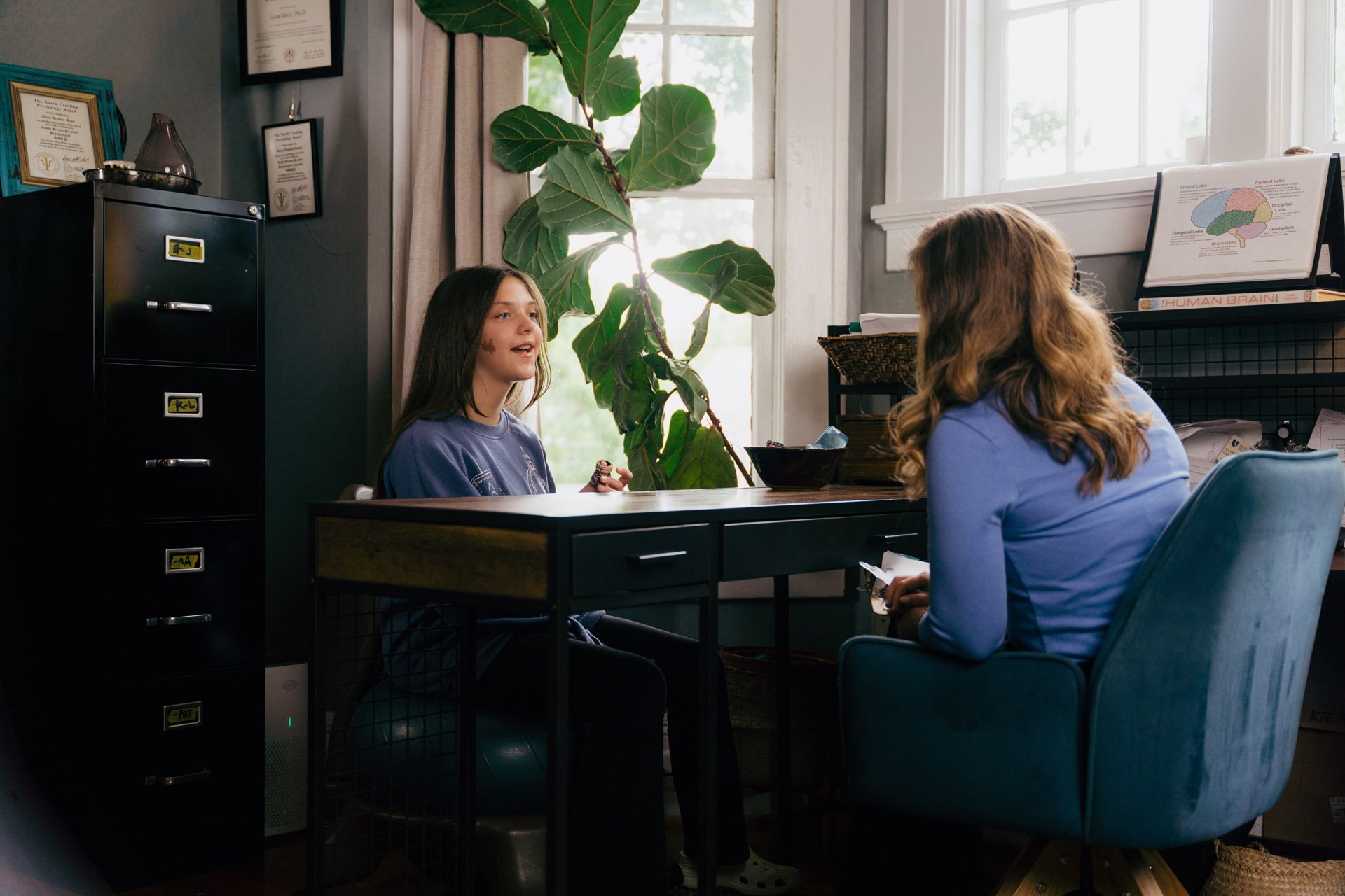A young girl and a woman are talking during a consultation in an office with a large green plant and educational posters on brain anatomy.