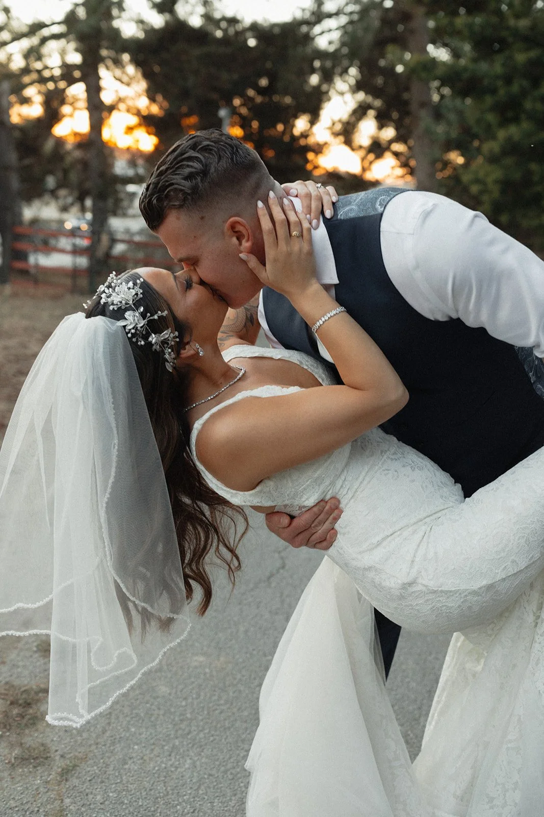 A groom passionately dips his bride for a romantic kiss at sunset. The bride wears a lace gown, veil, and pearl hairpiece, while the groom is dressed in a vest and white shirt. A magical wedding moment captured outdoors.