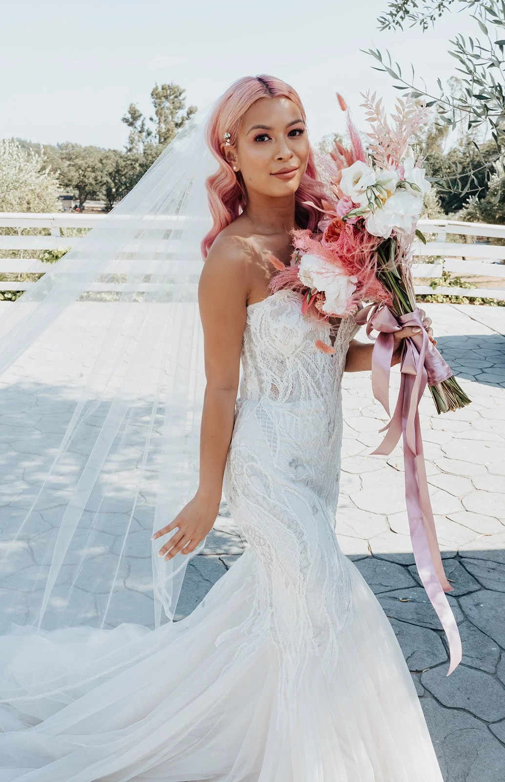 A bride with pastel pink hair poses in a white lace gown, holding a bouquet of pink and white flowers with flowing ribbons. Her veil billows behind her.