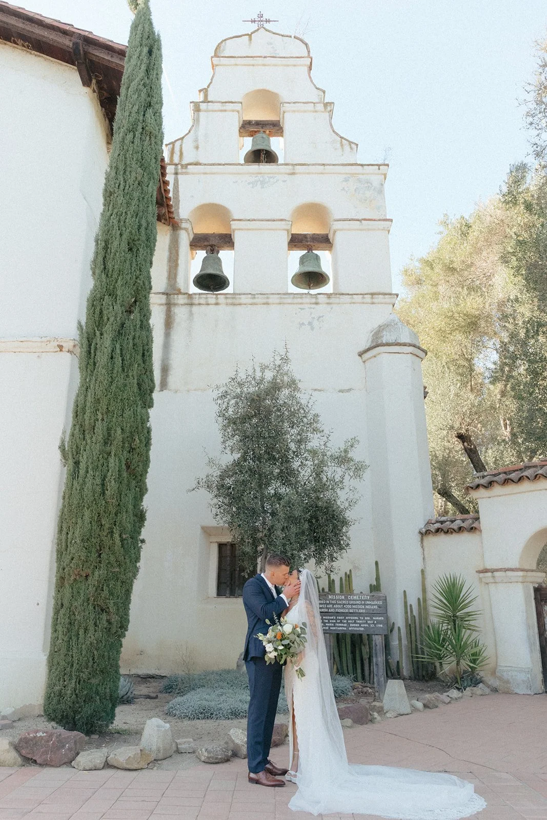 A newlywed couple shares a romantic kiss in front of a historic San Juan Batista Spanish-style mission with white stucco walls and bell towers. The groom wears a navy suit, and the bride wears a long veil and holds a bouquet.