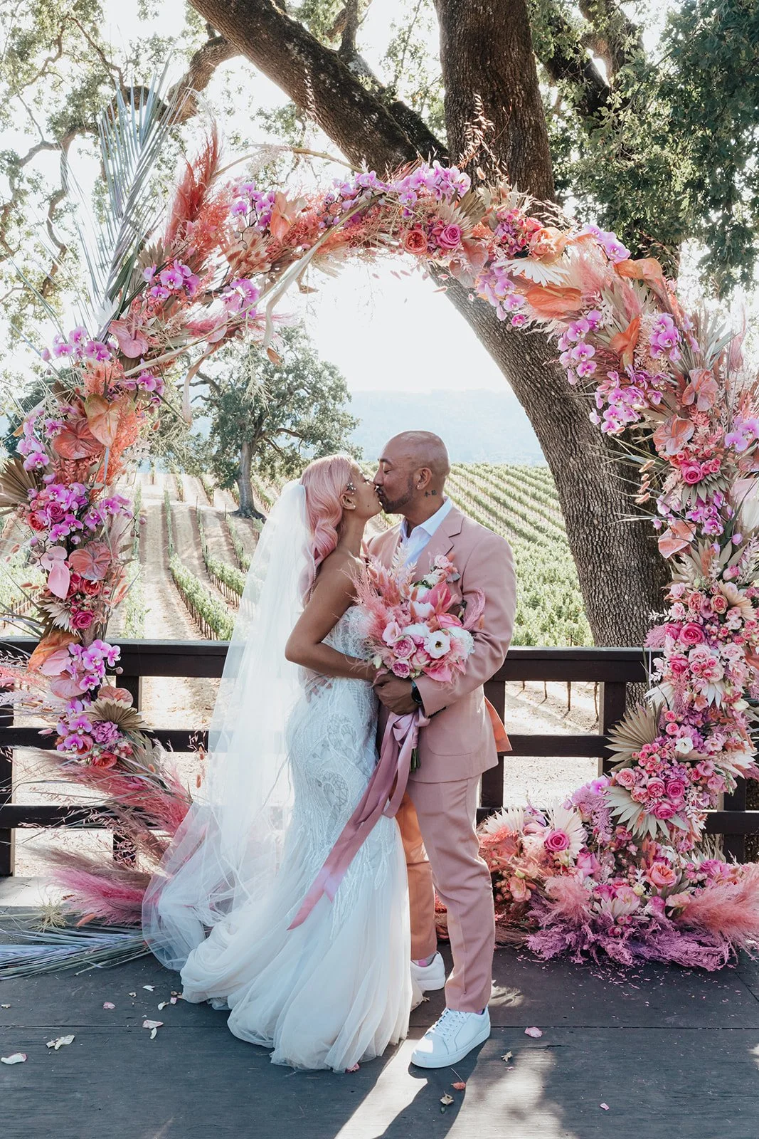 A newlywed couple shares a kiss in front of a pink floral arch with a vineyard backdrop. The bride wears a white gown and veil, while the groom wears a pink suit.