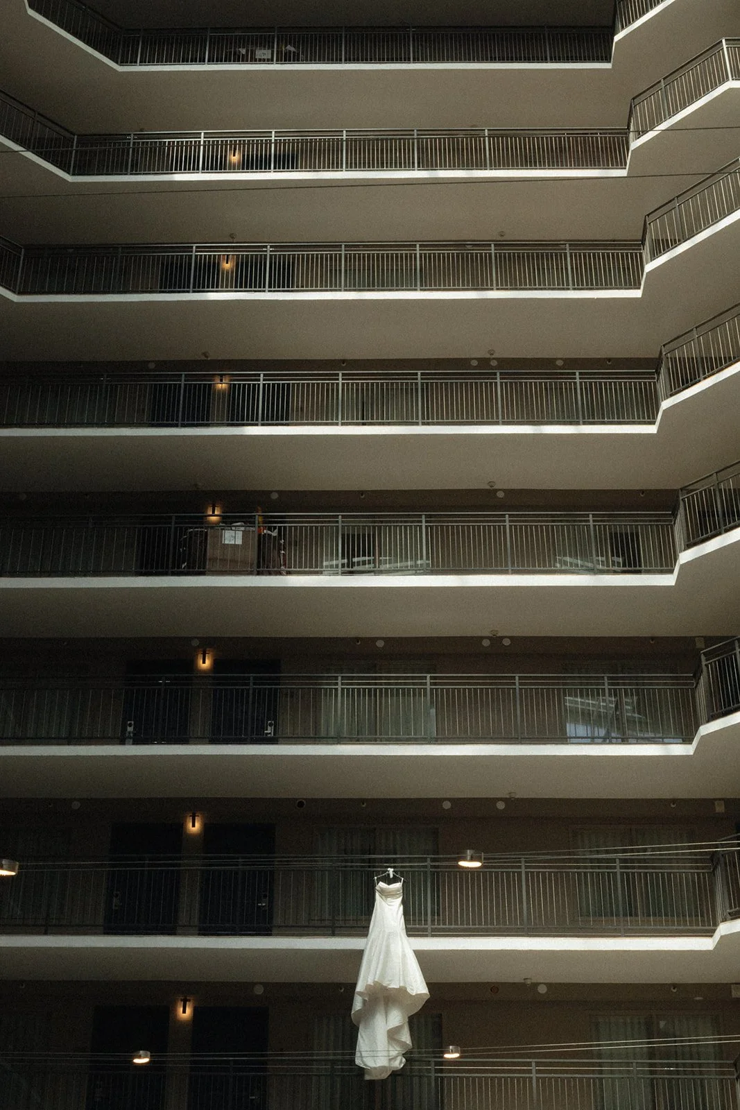 A wedding dress hangs elegantly in the center of a modern multi-story building, framed by symmetrical balconies. The dramatic composition highlights the gown’s beauty and anticipation of the big day.