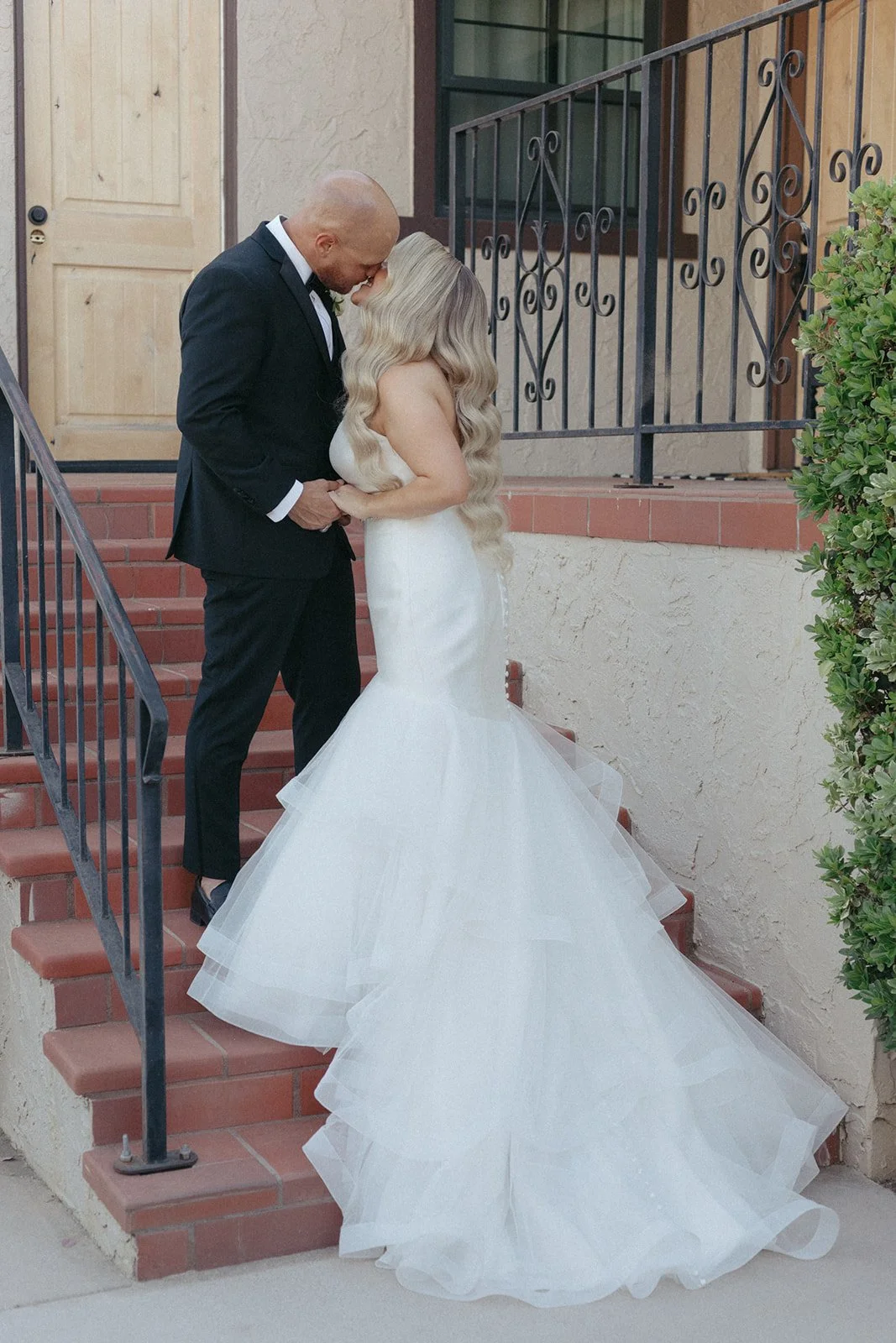 A bride and groom share an intimate moment on a brick staircase outside a Spanish-style building. The bride wears a tiered mermaid wedding gown, while the groom is dressed in a black suit. A romantic and elegant wedding portrait