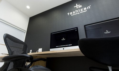 オフィス内のデスクとコンピューター、壁に「tashizan branding design」のロゴが表示。モダンなインテリア。