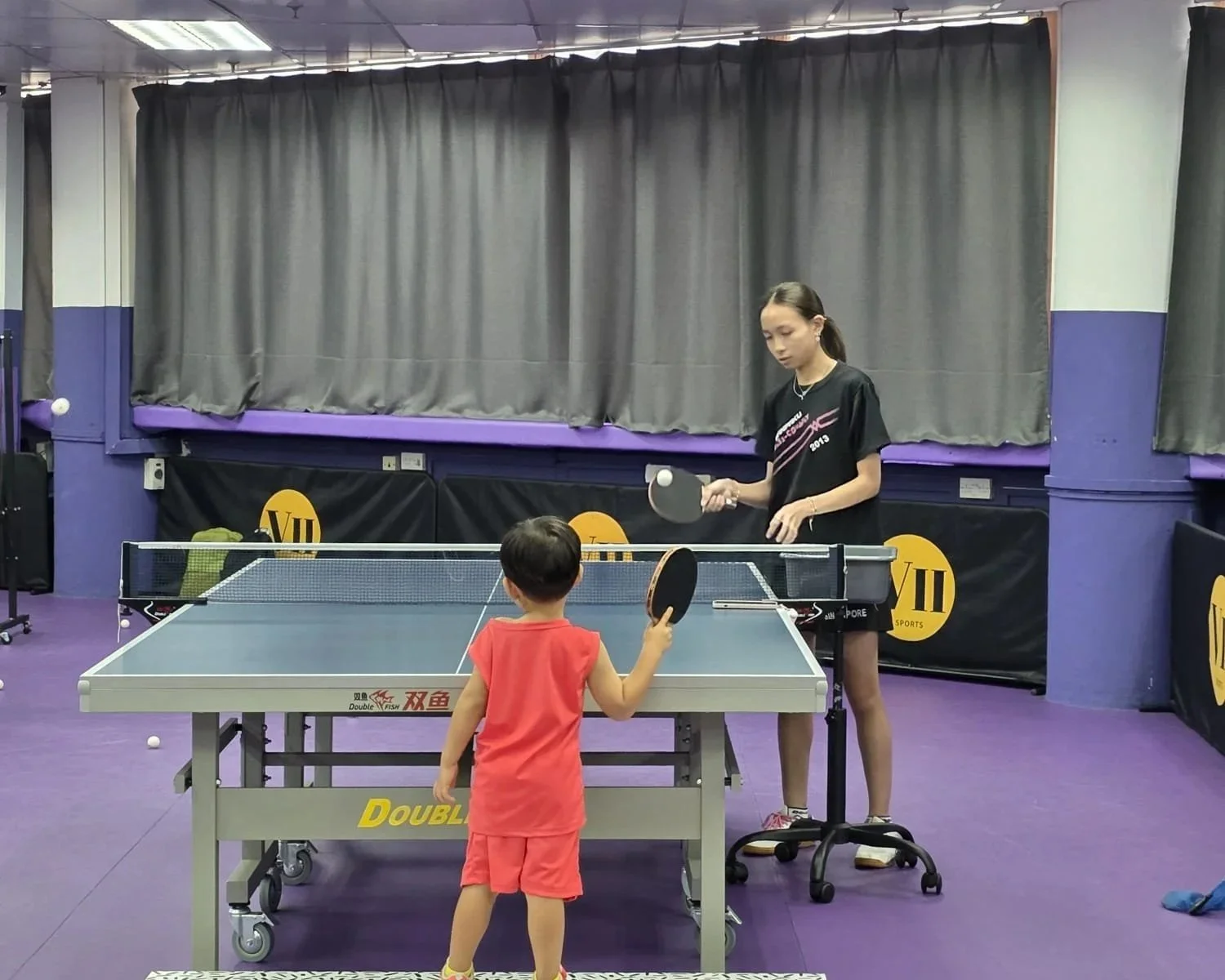 A young girl and a small child playing ping pong indoors, with the girl holding a paddle and the child reaching out towards the ball on the ping pong table.