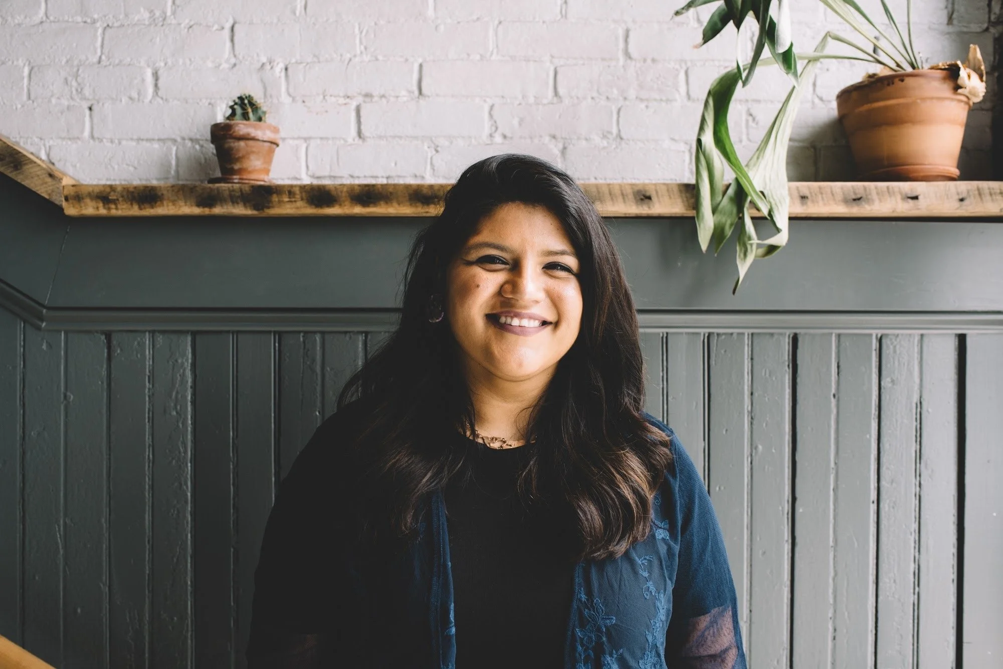A woman with long dark hair smiling in front of a white brick wall and a wooden shelf with potted plants.
