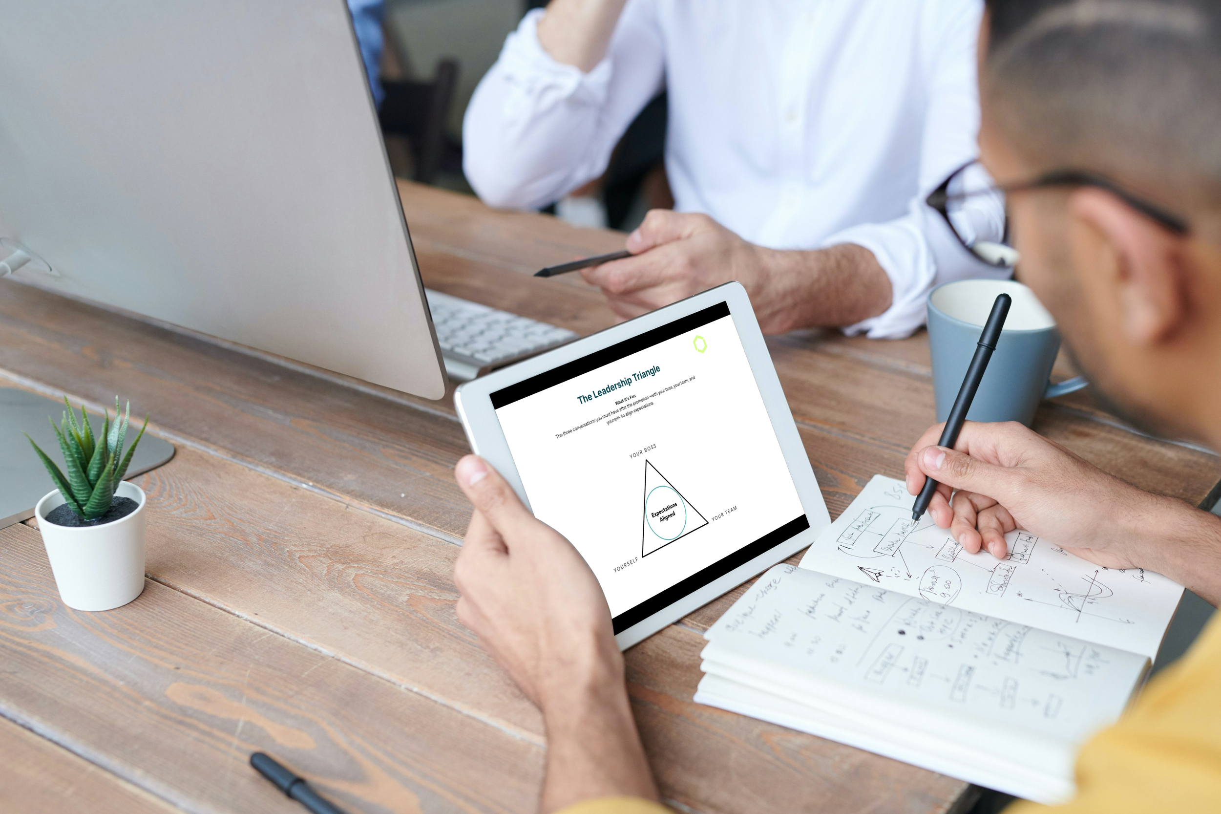 Two professionals in a modern office review a leadership framework diagram shaped like a triangle labeled "The Pyramid" on an iPad, with one taking notes—ideal for new managers building structured team strategies.