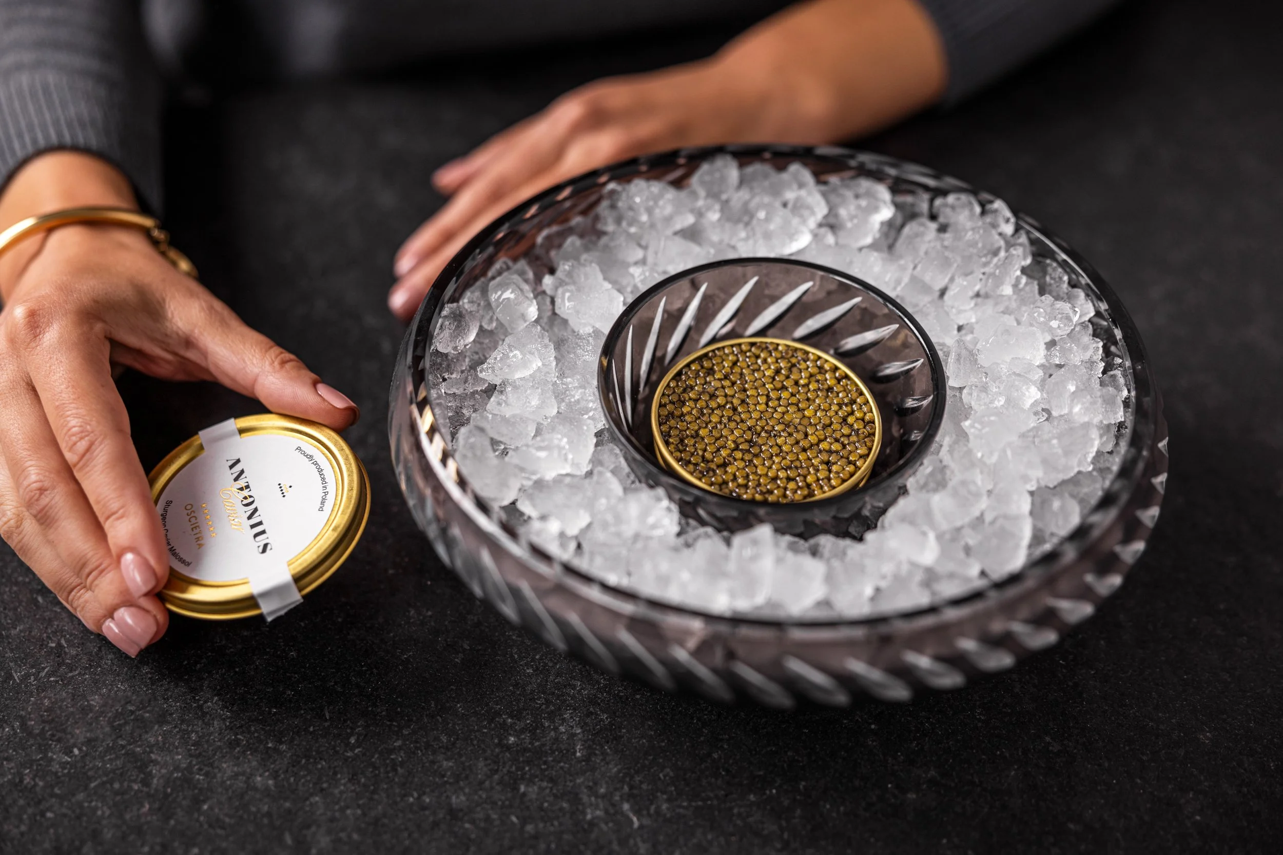 A person's hands holding a small can of smoked salmon near a decorative glass bowl filled with ice and a gold-colored container inside the bowl.
