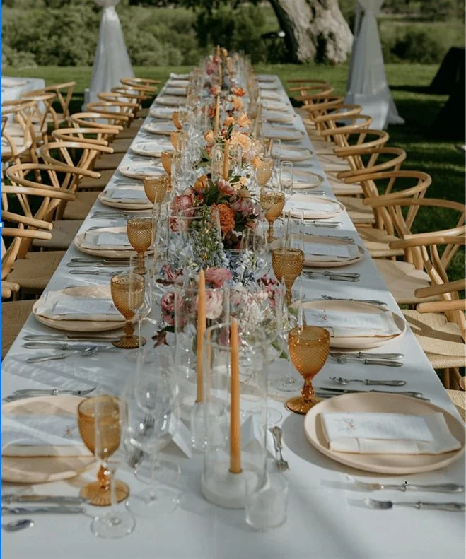 A long outdoor dining table set with floral centerpieces, candles, and elegant tableware, surrounded by wooden chairs on a grassy area.