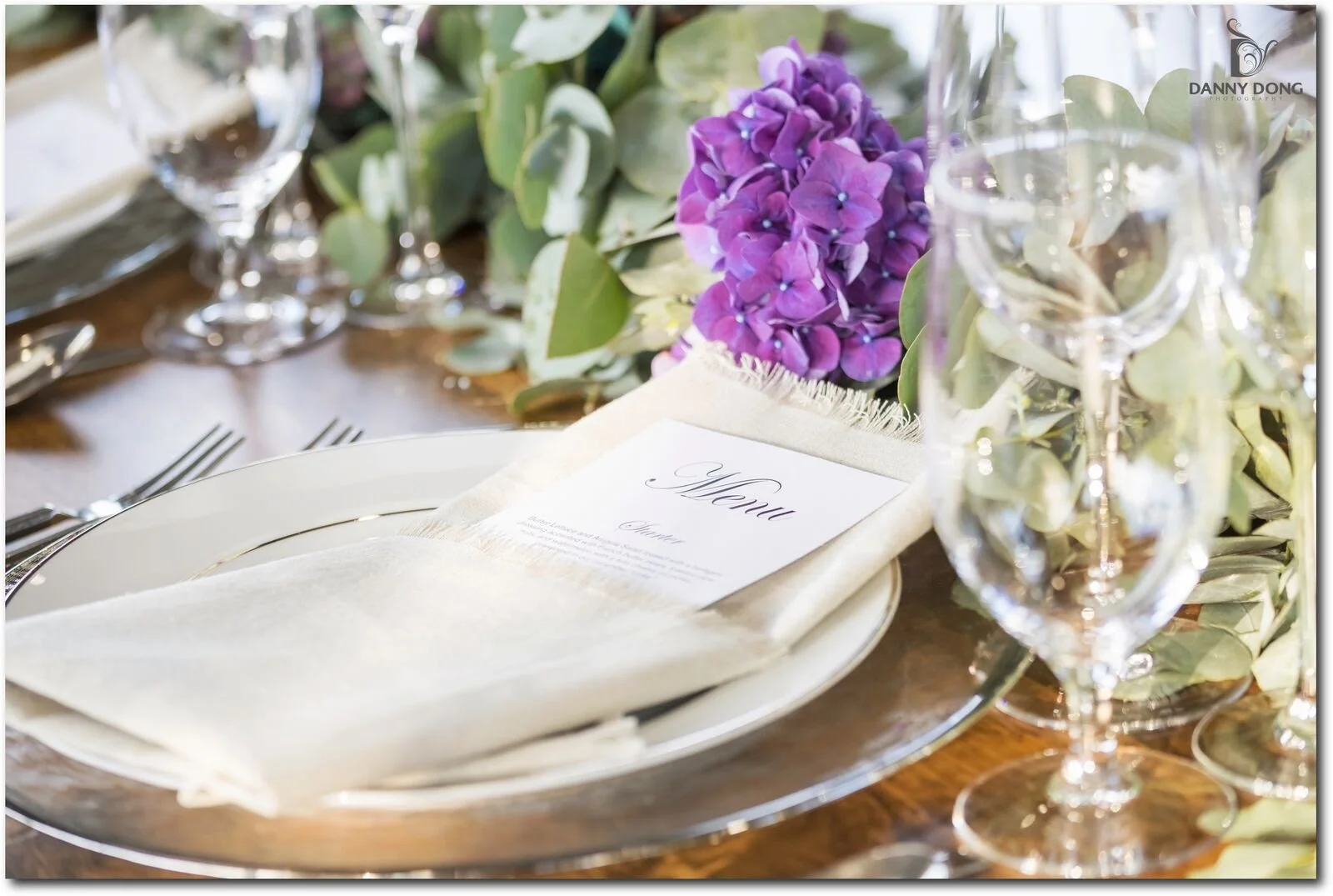 Elegant table setting with a menu on a napkin, purple flowers, greenery, and clear wine glasses on a wooden table.