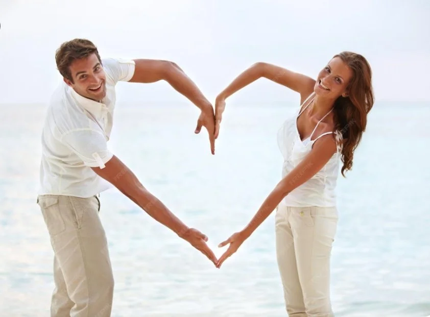 Smiling couple in white outfits forming a heart shape with their arms on a beach.