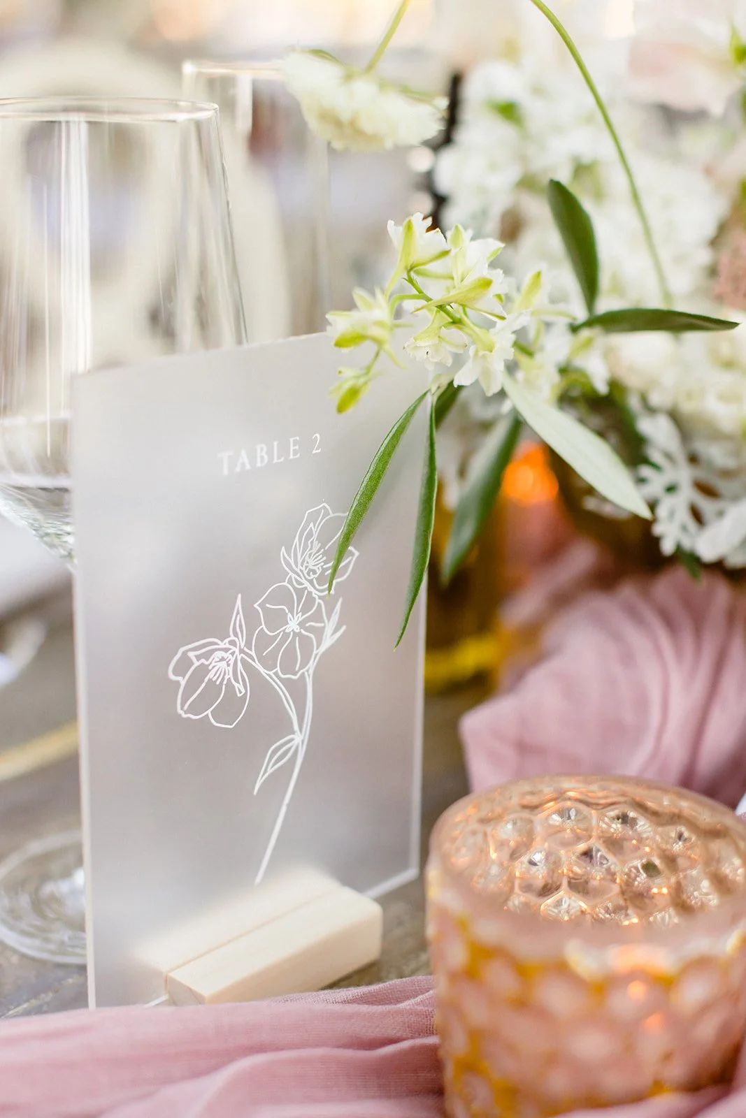 Table setup with a clear acrylic table number sign labeled "TABLE 2" decorated with white orchids, surrounded by glassware, floral arrangements, and a decorative pink candle holder.