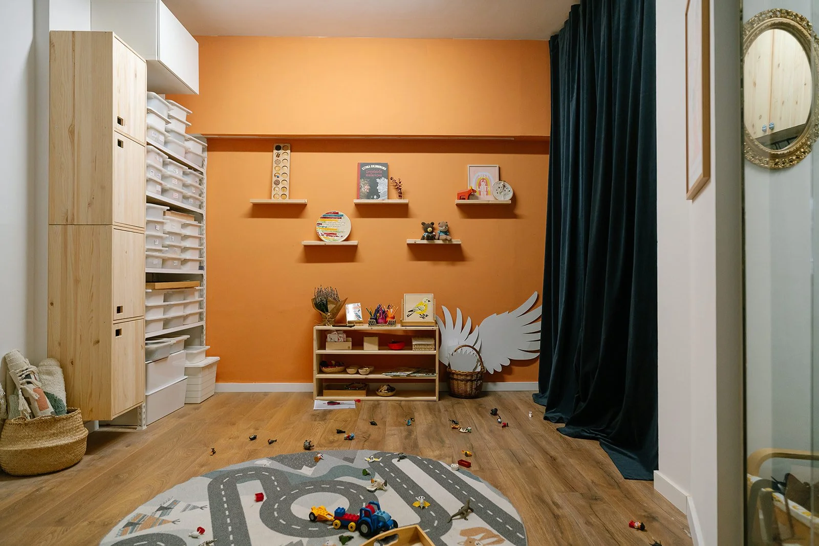 A children's playroom with a hardwood floor, orange wall, and dark blue curtain. Toys are scattered on the floor and a patterned rug with roads and vehicles. There are wall shelves with books and decorations, and a small white storage unit with toys and art supplies.