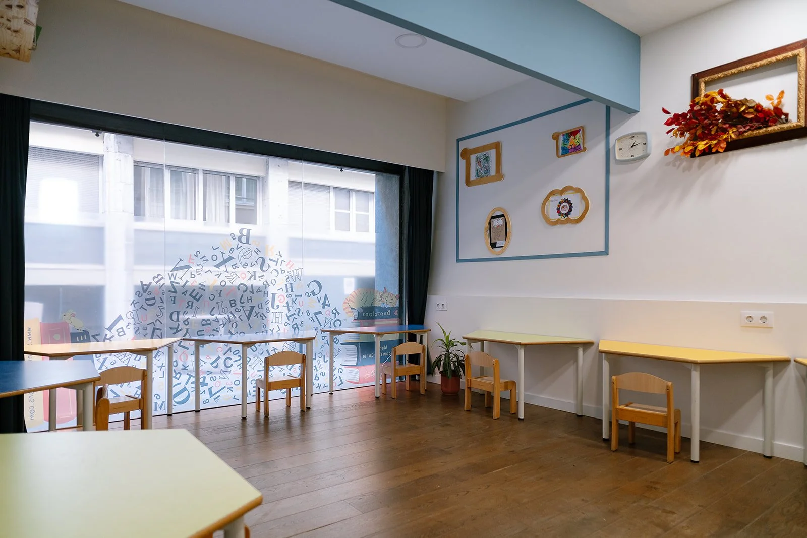 Empty classroom with small wooden tables and chairs, a large window with a decorative decal of scattered letters, a wall decorated with framed artwork, a clock, and a potted plant.
