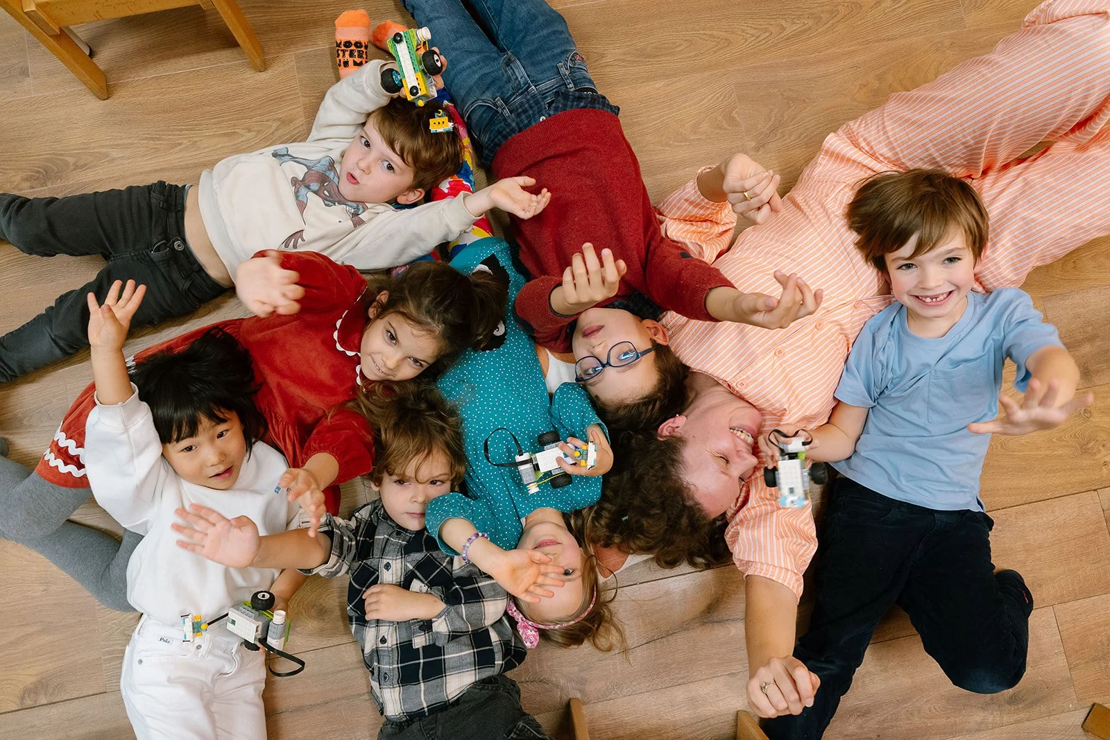 Grupo de niños y una mujer acostados en el suelo, jugando con pequeños robots y juguetes, algunos niños saludando y sonriendo.