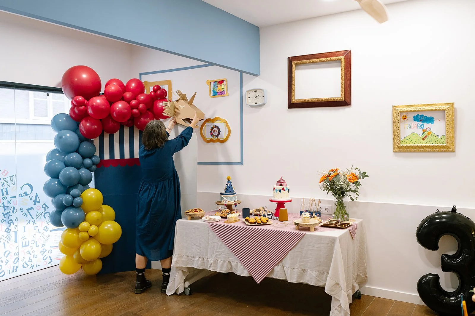 A woman in a blue dress decorating a birthday party with a gold unicorn balloon, nearby a table with birthday cakes and snacks, and colorful balloon arrangements in blue, red, and yellow, with framed children's drawings on the wall.