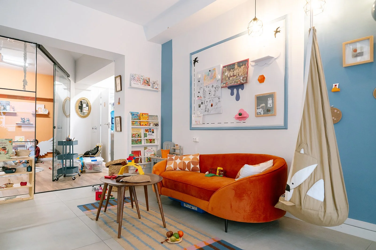 Children's playroom with orange velvet sofa, hanging fabric tent with animal face design, colorful artwork on the wall, small table with toys, bookshelves filled with children's books, and various toys scattered around.