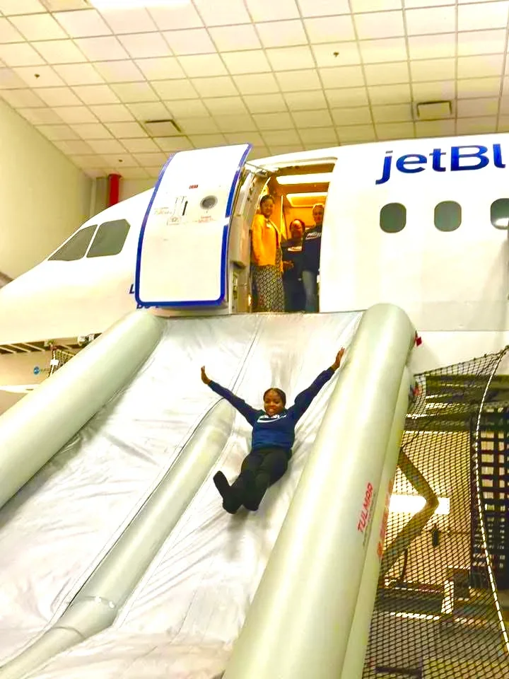 Child sliding down an inflatable slide at an indoor amusement park with a miniature airplane in the background.