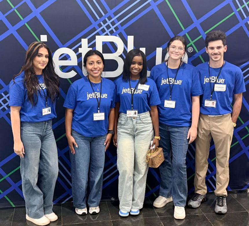 Group of five people wearing blue JetBlue shirts, standing in front of a JetBlue backdrop, smiling at the camera.