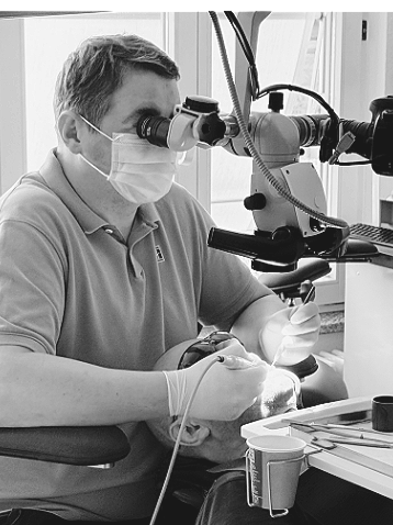 Dentist wearing magnifying glasses and a mask examining a patient's mouth using a dental microscope.