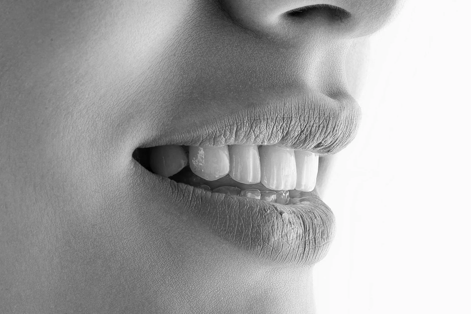 Close-up black and white photo of a person's lips and teeth, partially open, showing upper and lower teeth with textured skin surrounding the mouth.