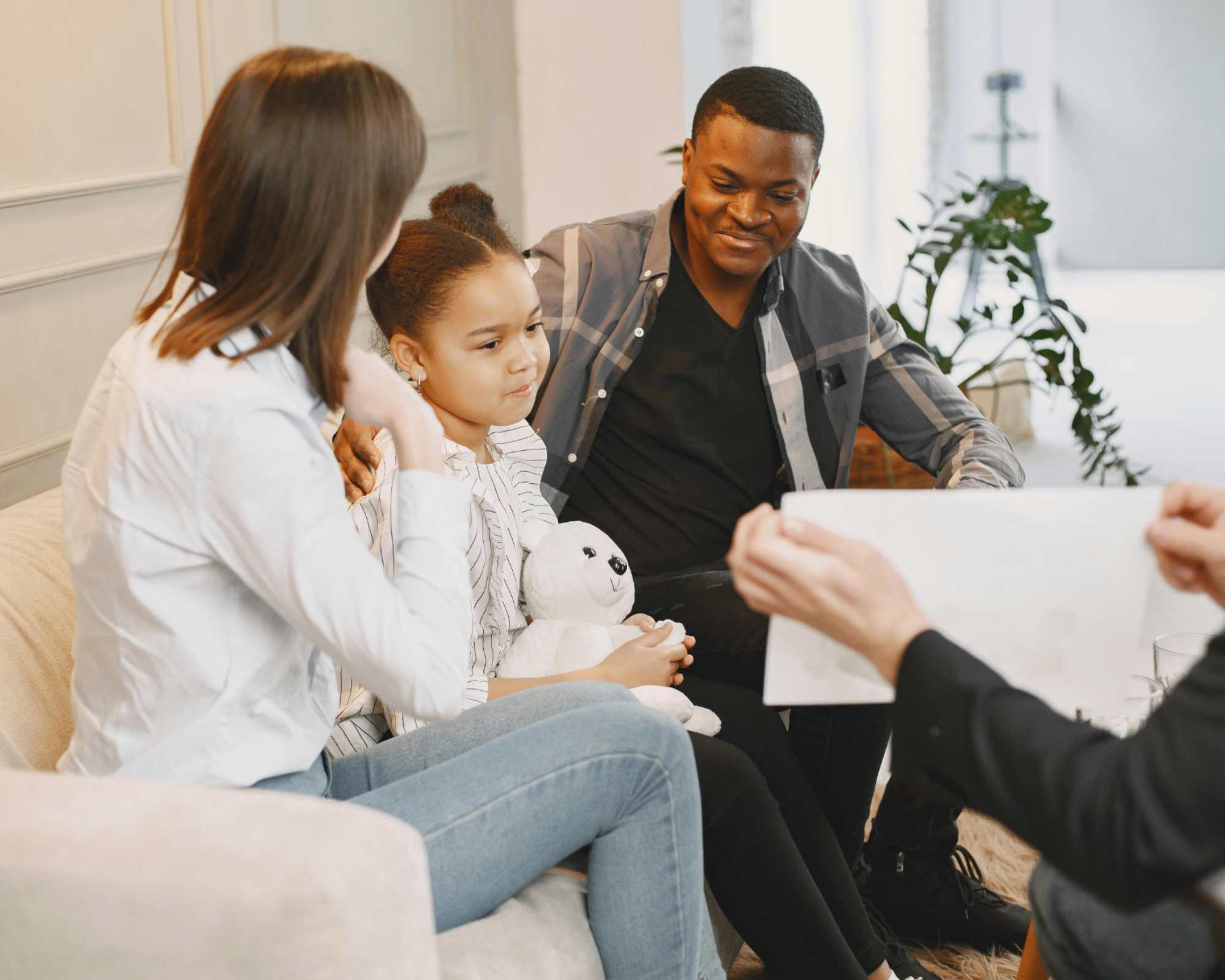 A happy family sitting on the floor indoors, with a smiling toddler in the center. The mother is on the left wearing a white sweater, and the father is on the right in a blue shirt. They are surrounded by toys.