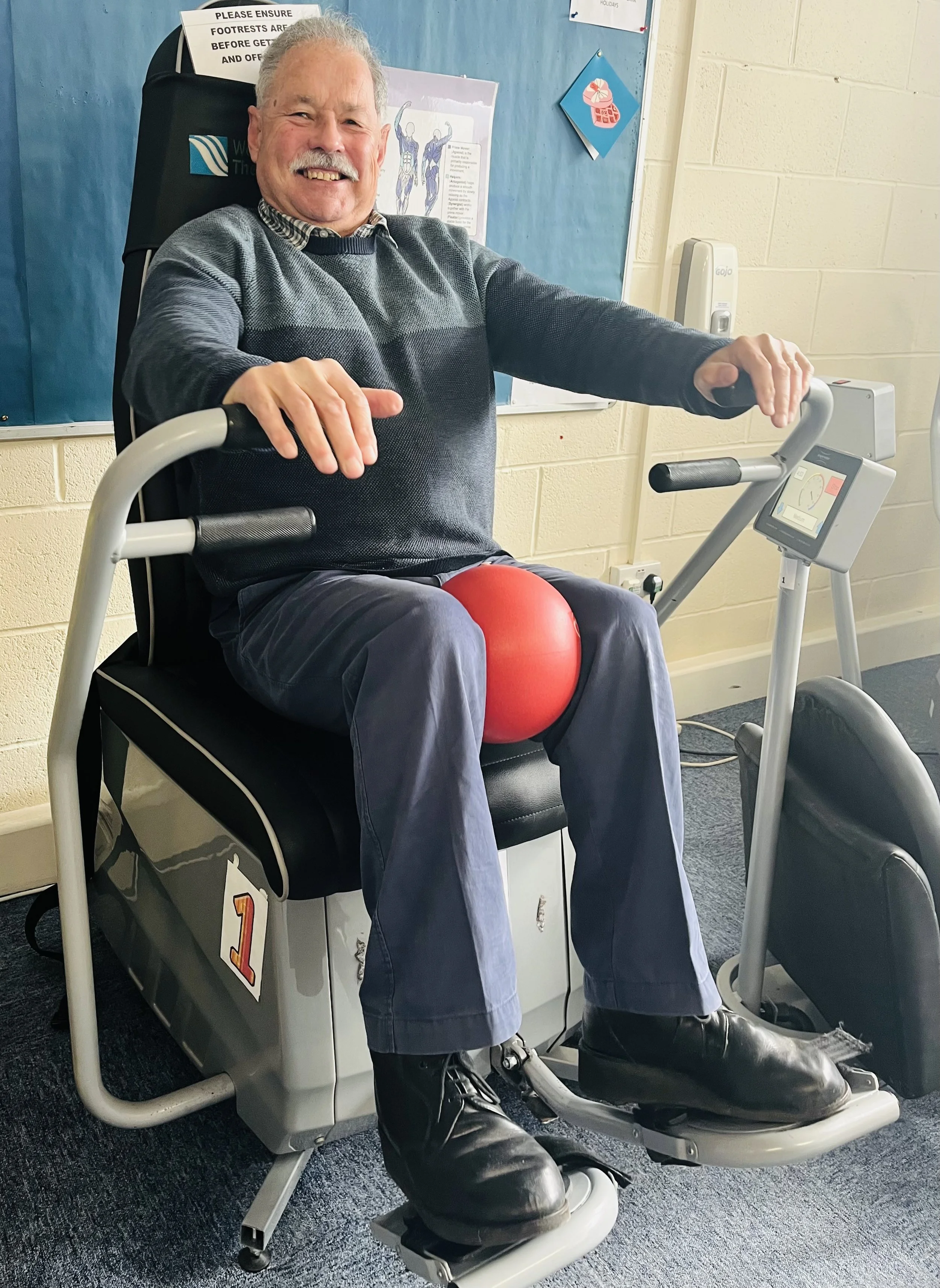 An elderly man sitting in a rehabilitation exercise machine with a red ball, smiling, in a clinical setting.