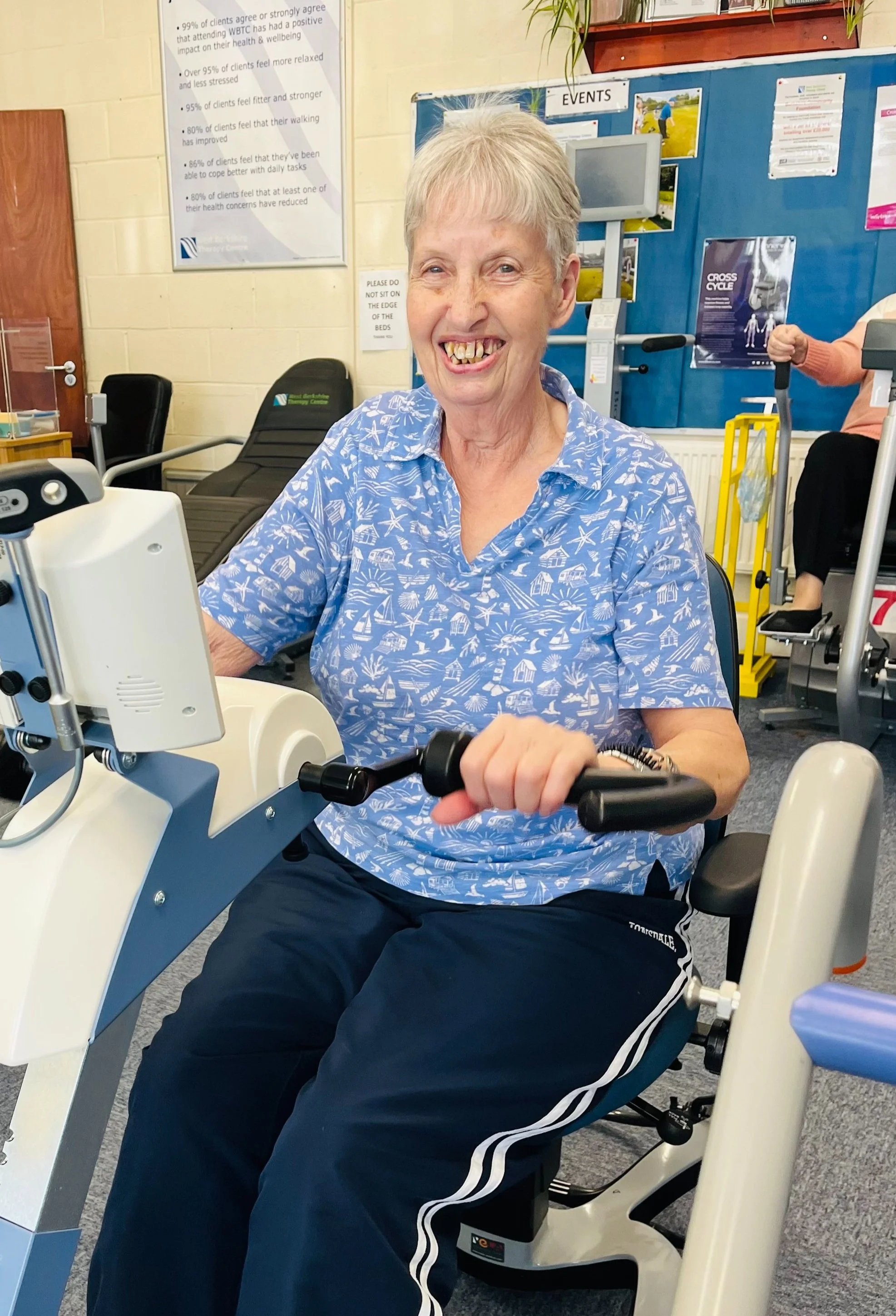 An elderly woman with short gray hair and wearing a blue shirt with white beach and boat patterns, smiling while sitting on a recumbent exercise bike in a gym or physiotherapy center.